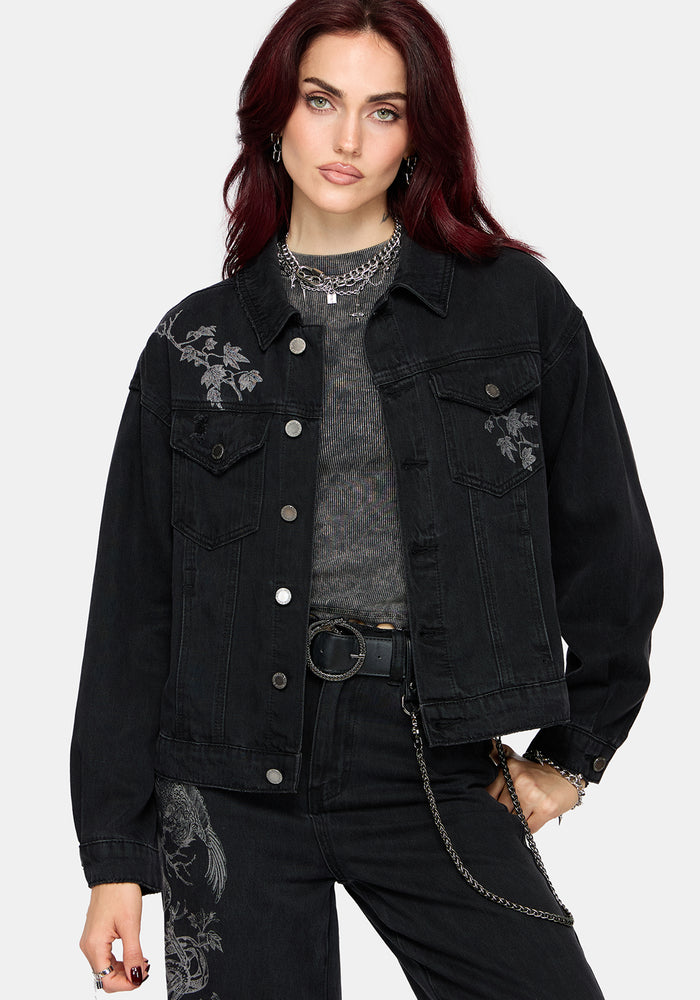 A model with dark red hair wearing a black denim jacket with grey floral embroidery on the chest, layered over a gray textured top and black jeans.