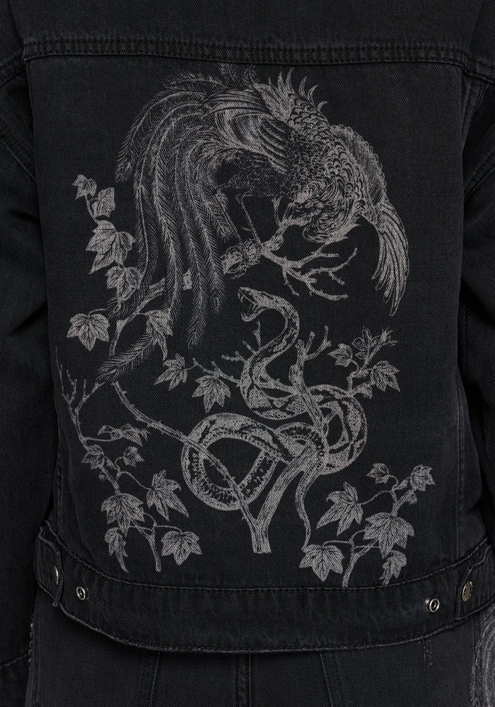Close-up of the back of a black denim jacket featuring a large grey embroidered graphic of a bird perched on a tree with a snake and leaves.