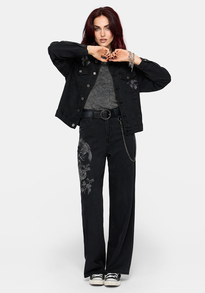 A model wearing a black denim jacket with floral embroidery, a grey knit top, black wide-leg jeans with matching embroidery, a black belt, and black sneakers, posed against a light gray studio background.