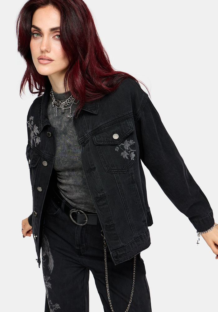 A model with dark red hair wearing a black denim jacket with floral embroidery, styled over a gray t-shirt and black pants against a light gray background.