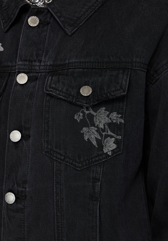 Close-up of a black denim jacket featuring a chest pocket with a gray embroidered leaf pattern and silver-toned metal buttons.